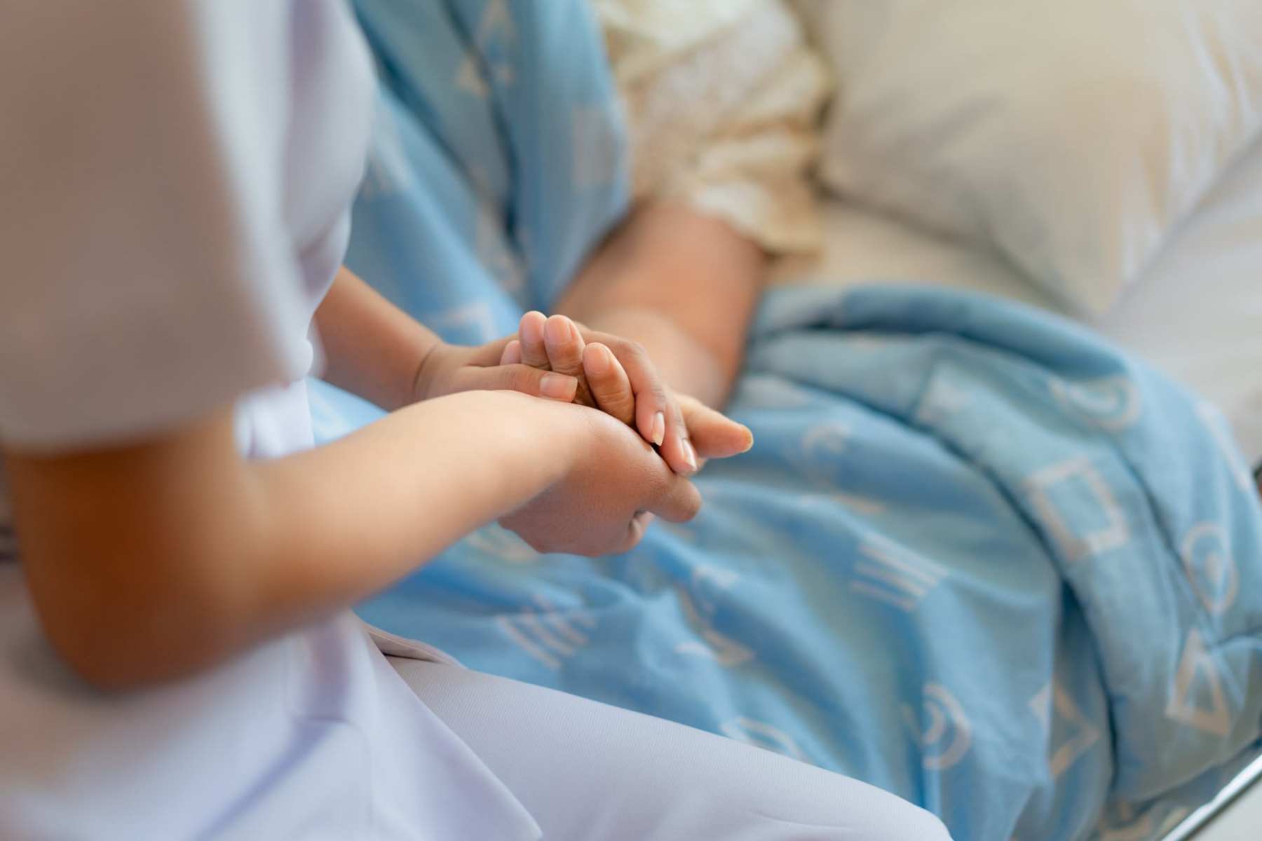 A nurse sitting on a hospital bed next to an older woman holding her hand.