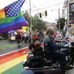 A person on a motorcycle with a rainbow flag attached to the back drives by protesters with a Trump-Pence sign.
