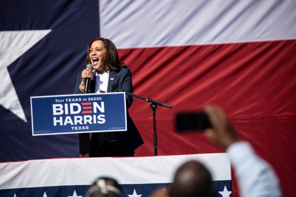 Kamala Harris stands at a podium at a campaign event with a giant Texas flag behind her.