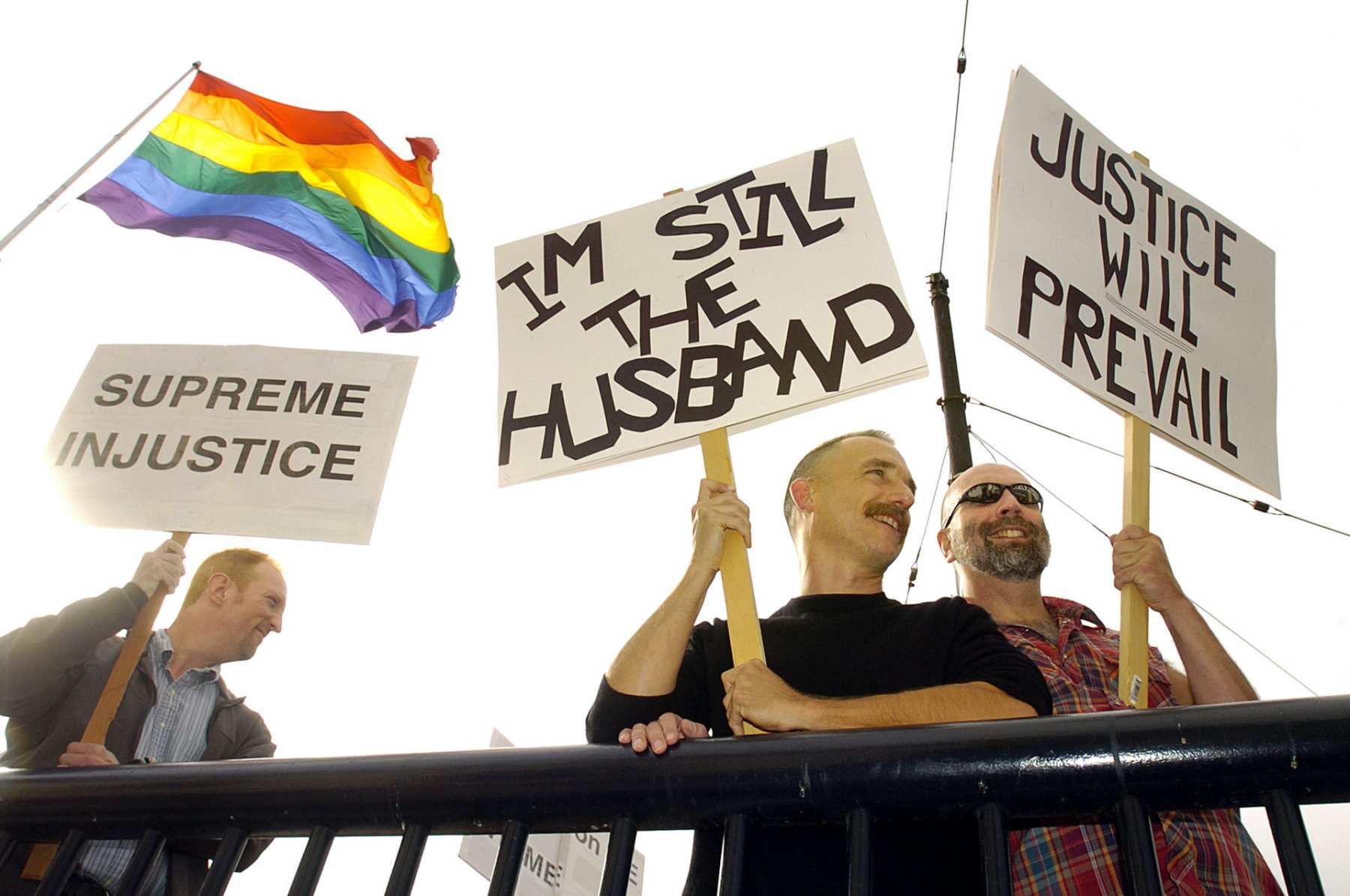 Three people stand with signs supporting marriage equality as the LGBTQ flag waves in the background.