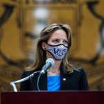 Michigan Secretary of State Jocelyn Benson stands at a podium with a mask on that says 