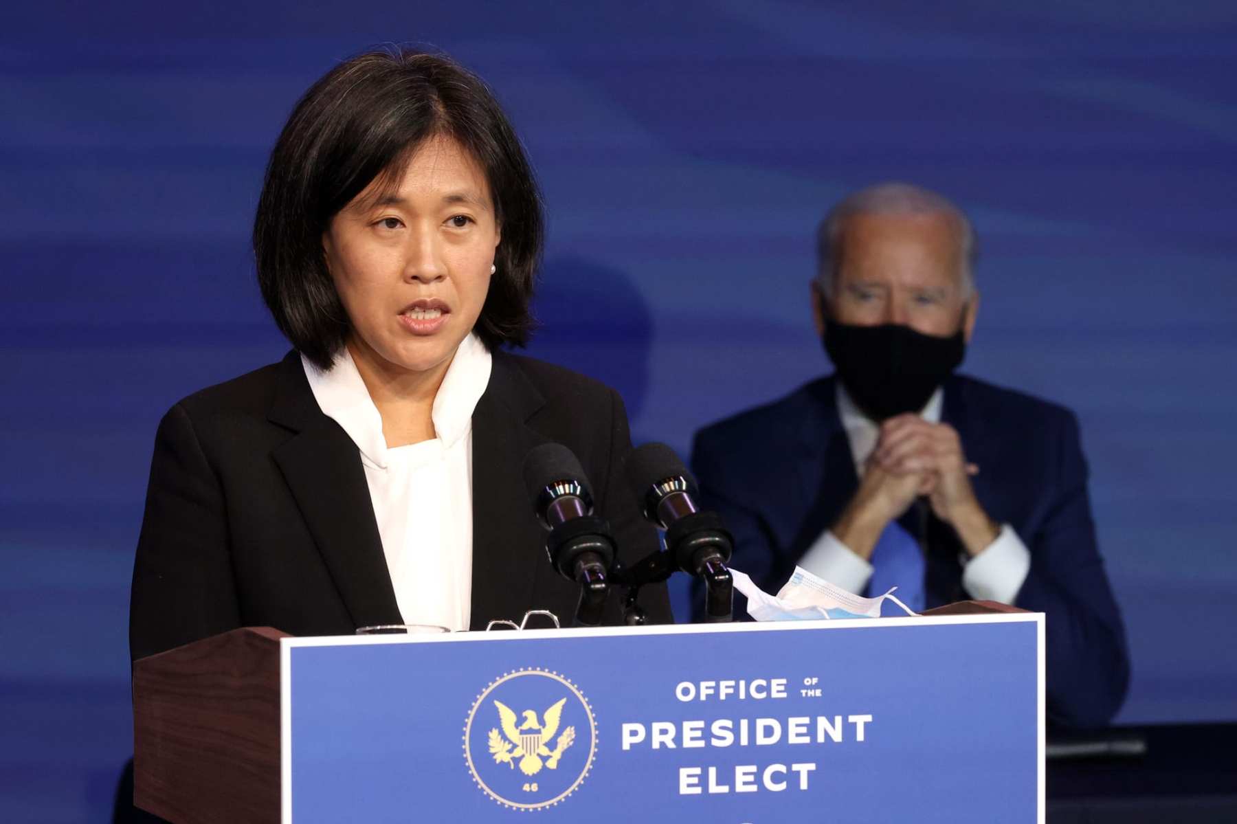 Katherine Tai delivers remarks from a podium with a masked Joe Biden standing behind her.