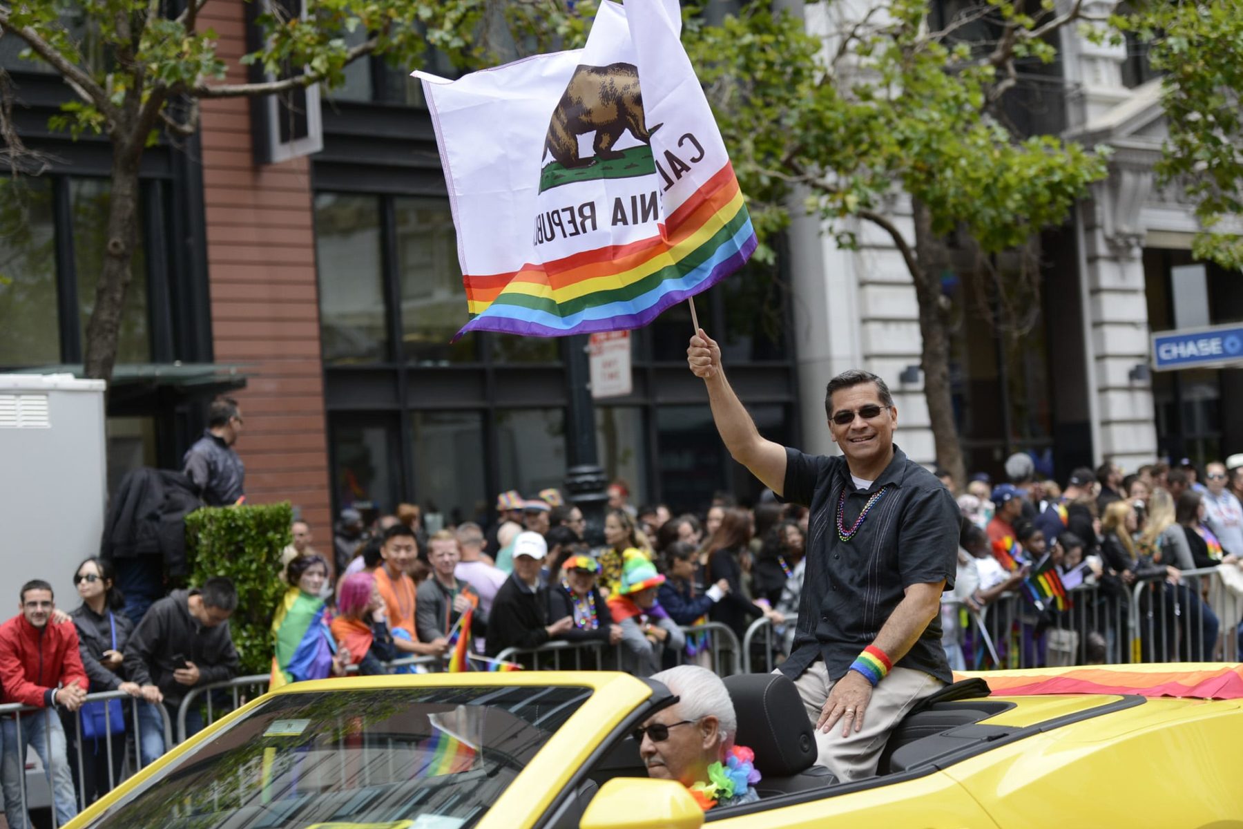 Xavier Becerra riding in a car waving a California flag with the LGBTQ+ rainbow at the bottom.