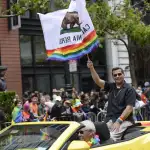 Xavier Becerra riding in a car waving a California flag with the LGBTQ+ rainbow at the bottom.