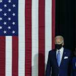 Joe Biden and Kamala Harris, both wearing masks, walk in front of an American flag.