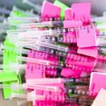 Unused doses of the COVID vaccine in a pile with bright pink and green labels.