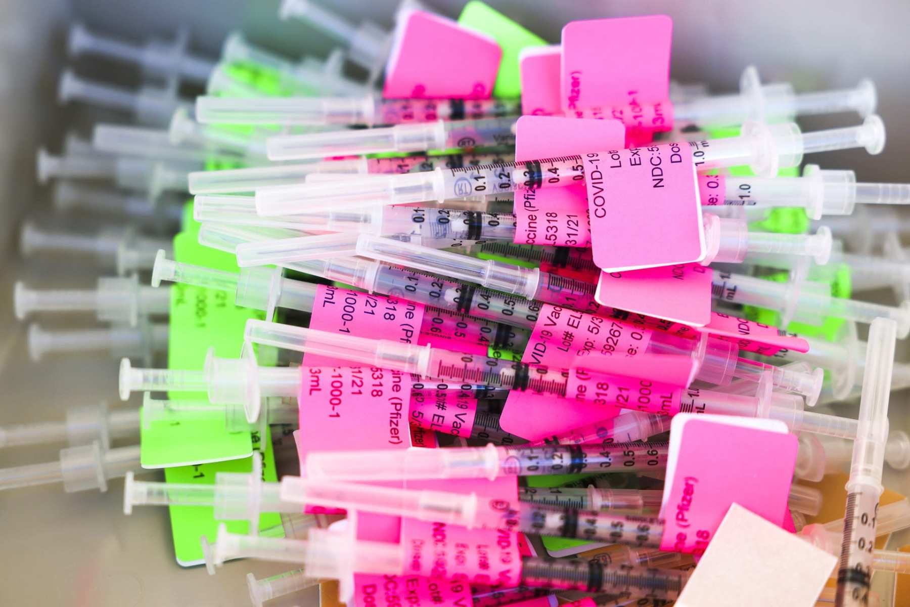 Unused doses of the COVID vaccine in a pile with bright pink and green labels.