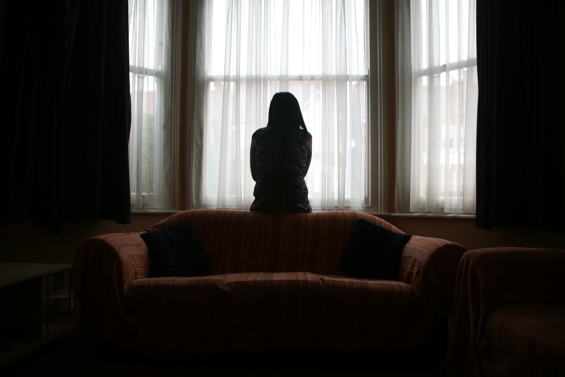 A young Asian woman suffering from domestic violence stands alone in the bay window of her home.
