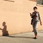 A woman walking down a street in NYC with a mask on looking at her phone.