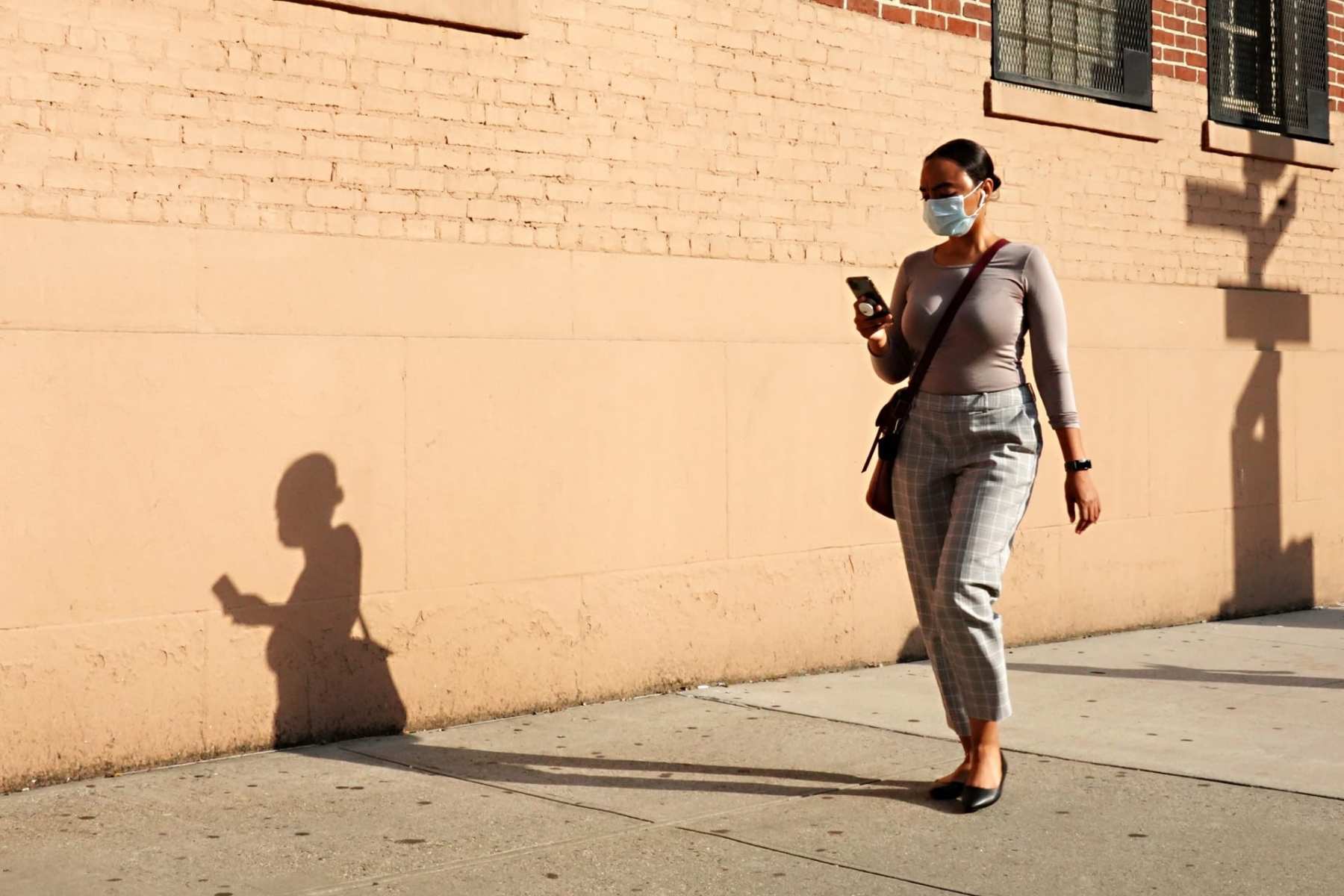 A woman walking down a street in NYC with a mask on looking at her phone.