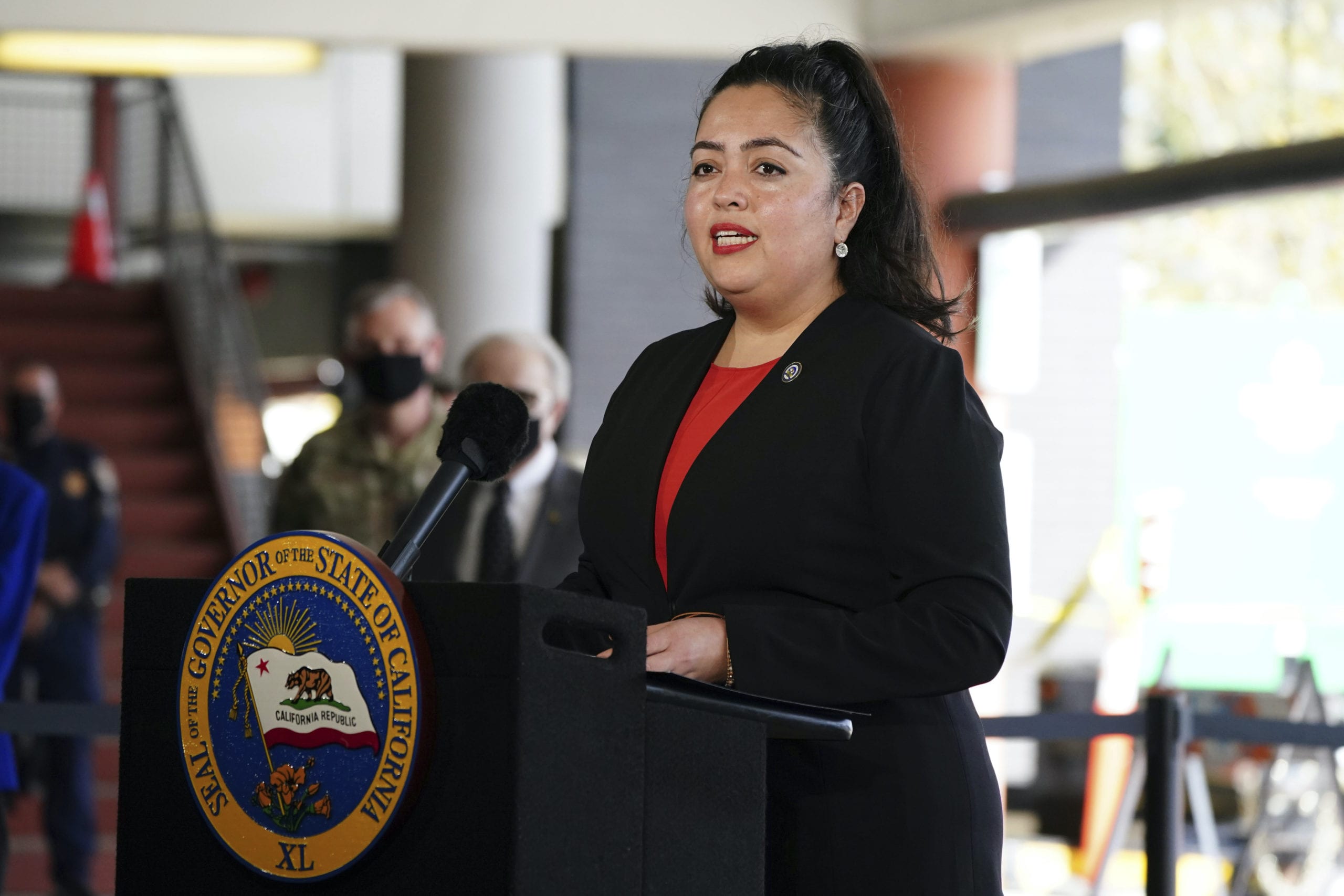 California Assemblywoman Wendy Carrillo (D-Los Angeles) stands at a podium in a red blouse and dark blazer speaking during a news conference at a joint state and federal COVID-19 vaccination site set up on the campus of Cal State LA.