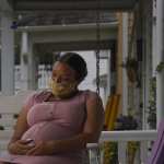 A pregnant woman sits on a porch swing holding her belly with a mask on.