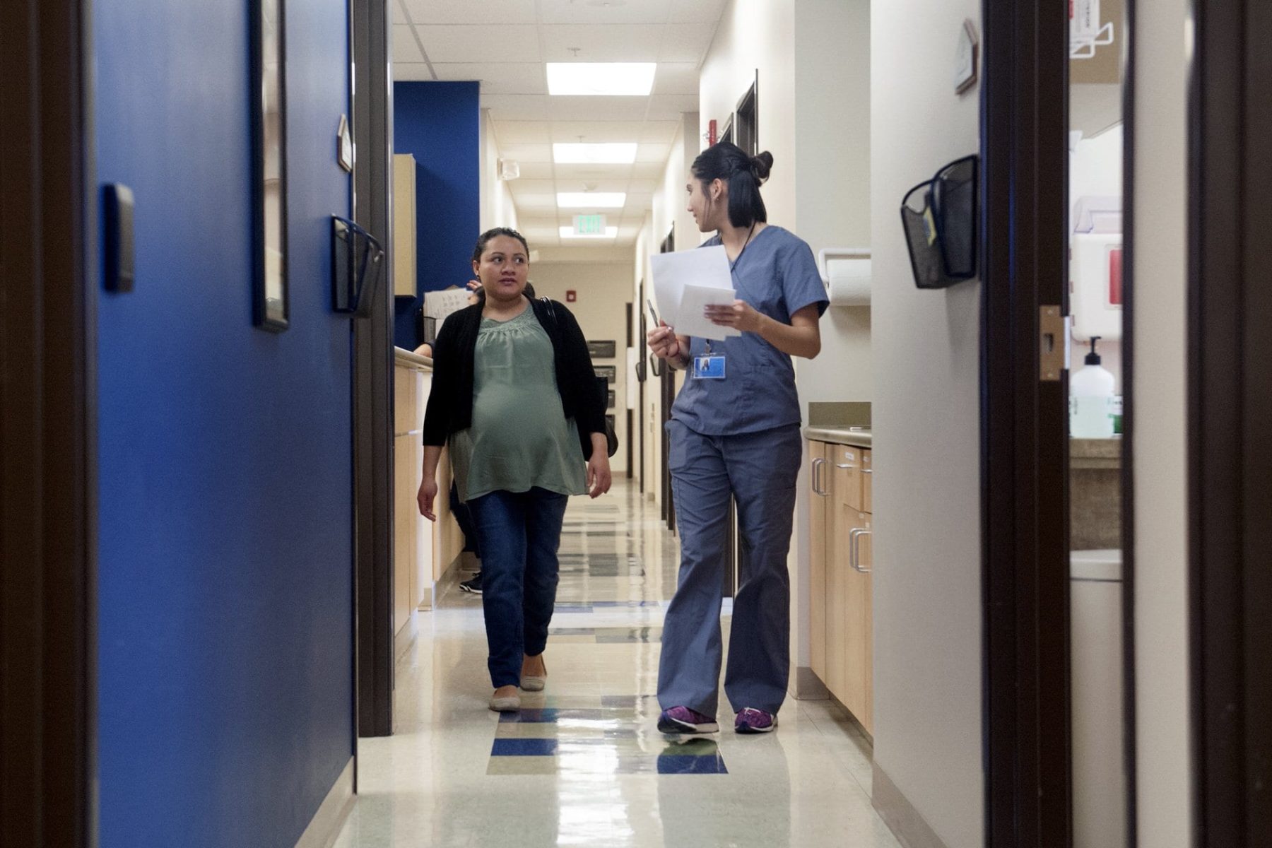 A pregnant woman walks down the hallway of a medical facility with someone in scrubs.
