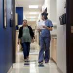 A pregnant woman walks down the hallway of a medical facility with someone in scrubs.