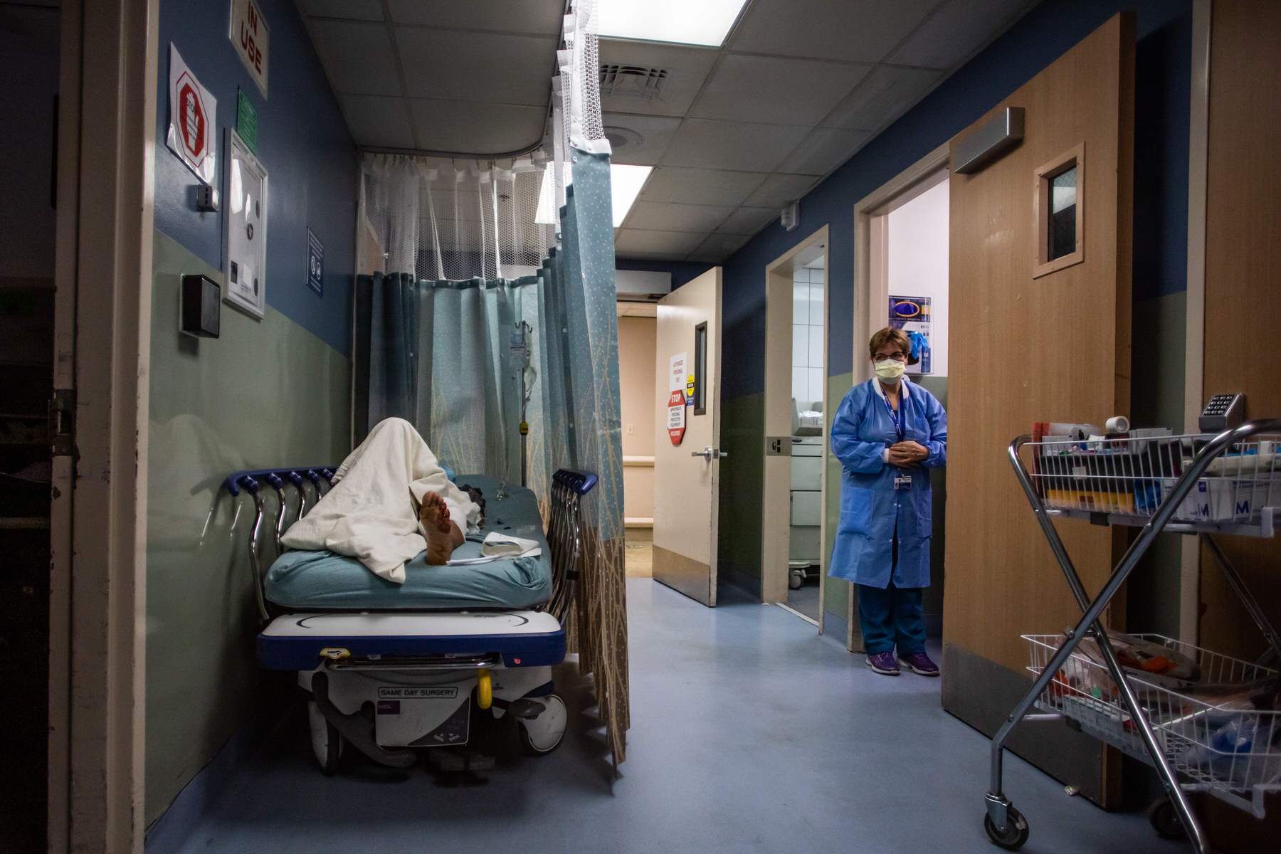 A patient rests in a corridor as a nurse looks on in the distance.