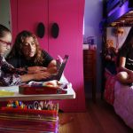 Two girls sit in their room on laptops during remote learning as mom helps.