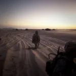 U.S. soldiers kneel in the sand looking at tanks in the distance at sunset.