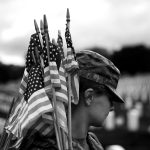 A female soldier holds American flags over her shoulder. Her face is partly obscured by her cap.