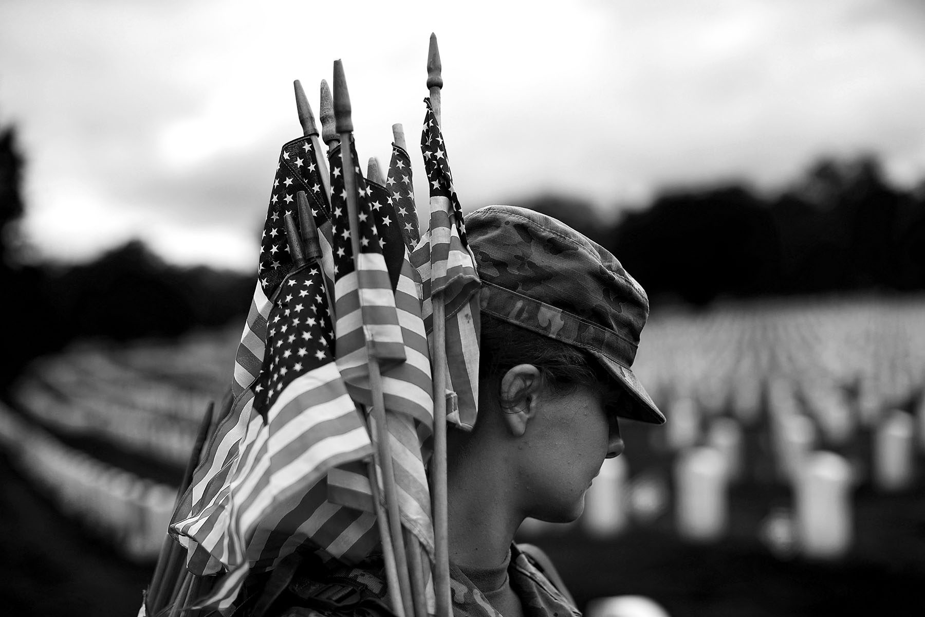 A female soldier holds American flags over her shoulder. Her face is partly obscured by her cap.