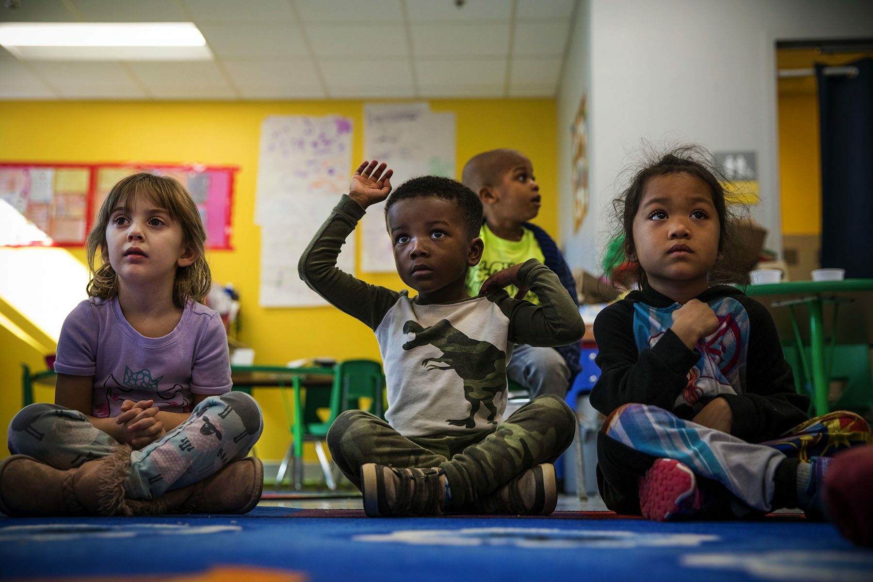 Three small children are seen in a colorful classroom. The child in the middle raises his hand while the other two appear to be listening..