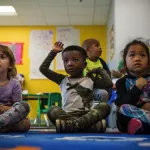 Three small children are seen in a colorful classroom. The child in the middle raises his hand while the other two appear to be listening..