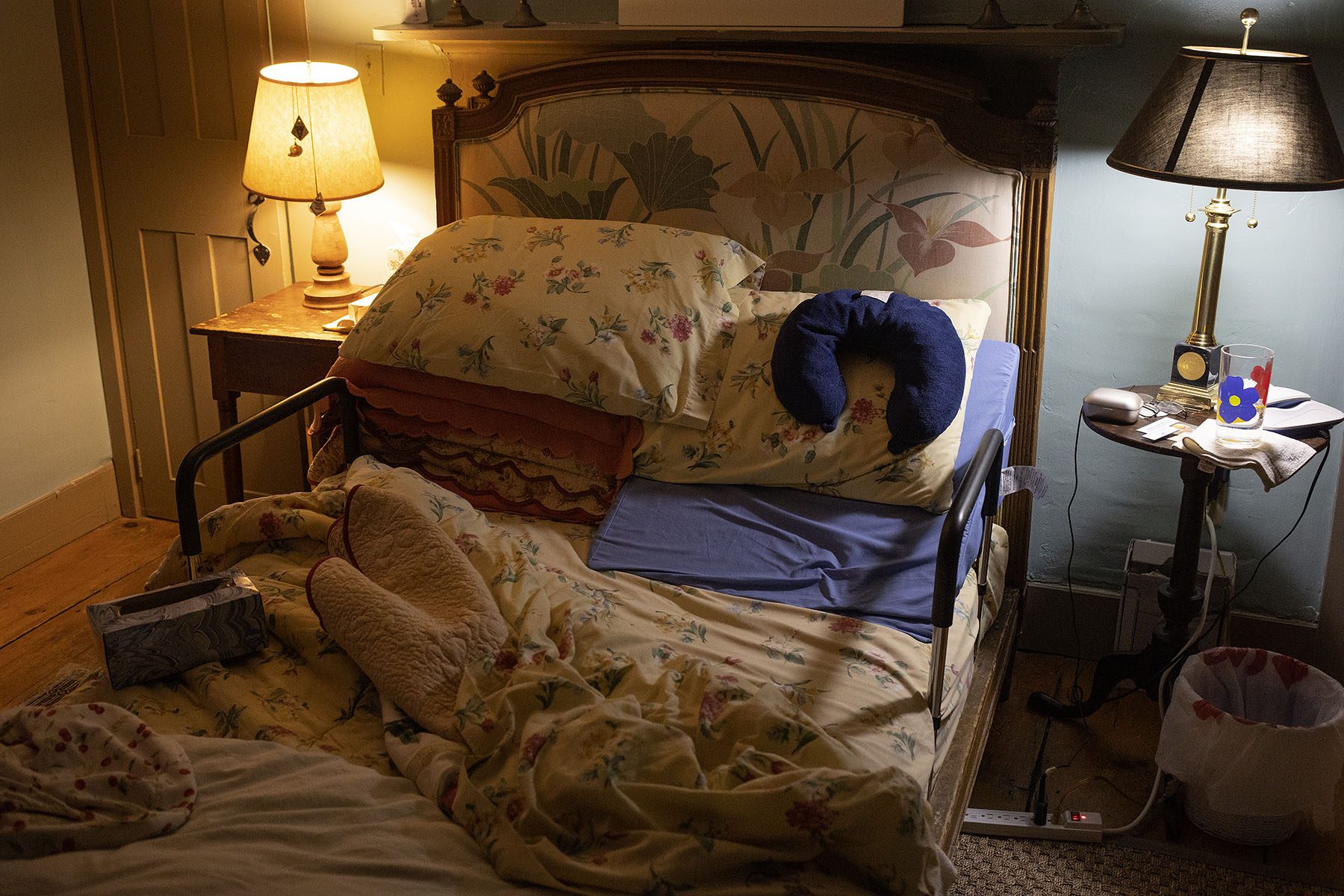 The bedroom of an elderly person is pictured. The bedsheets are undone and on the bed rests a neck pillow and a box of tissue paper that appears empty.