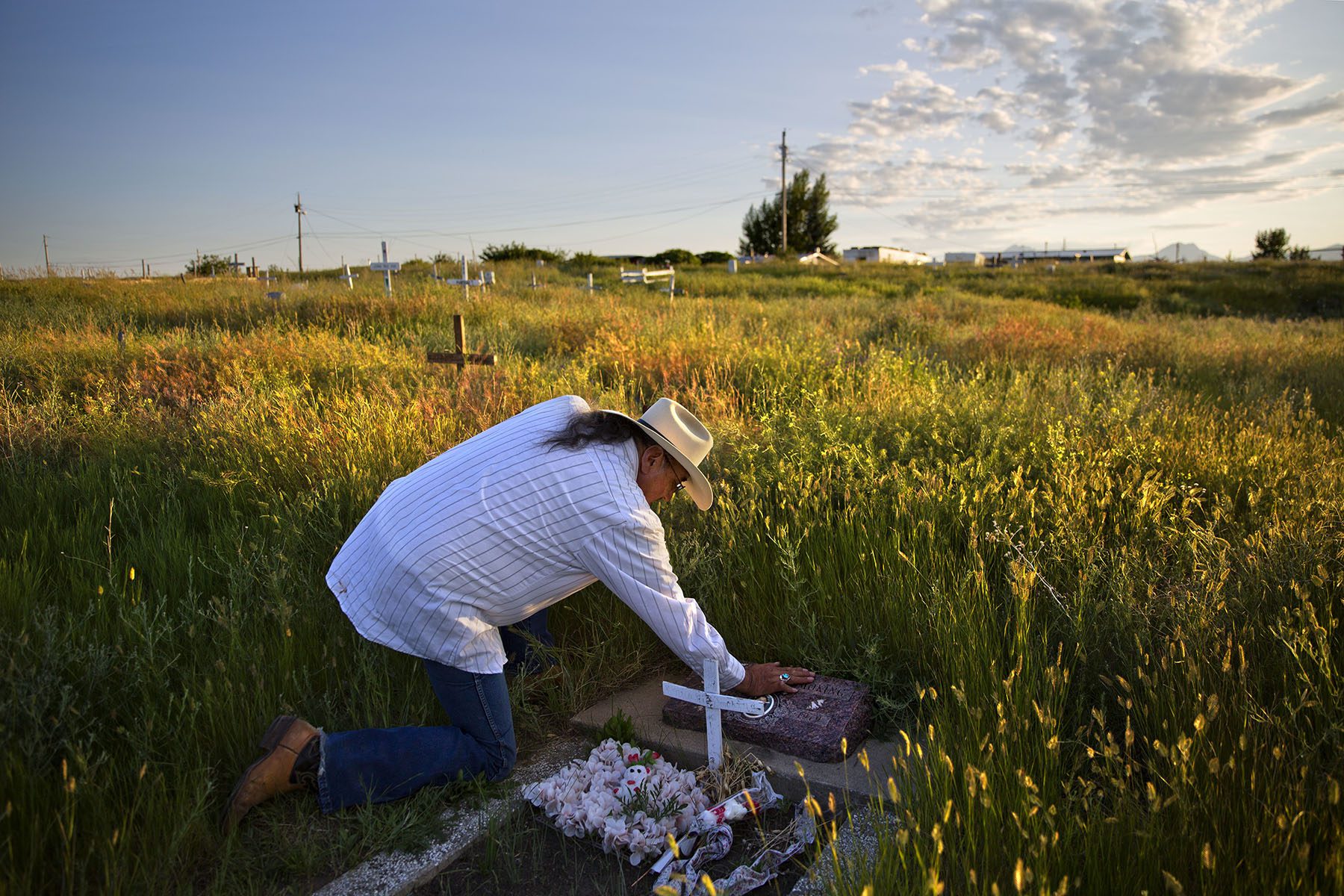 A man wearing a cowboy hat kneels to touch a tomstone in a field at golden hour.