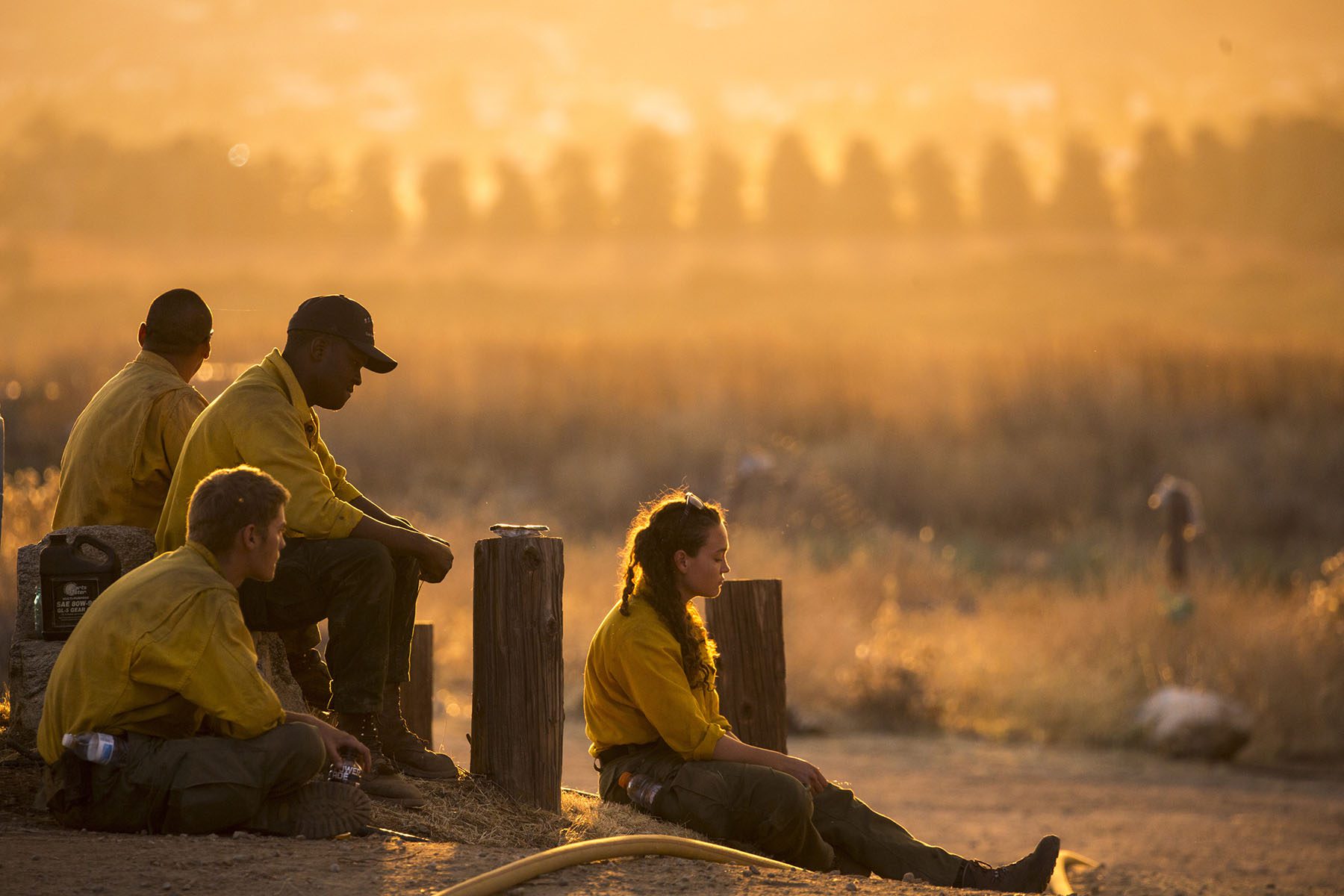 Three male firefighters and one female sit on the ground as golden light and smoke is seen in the distance.