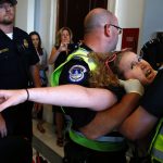 A woman is being carried by multiple capitol police officers. She seems to be struggling greatly.