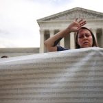 A protester waves her arm into the sky and closes her eyes while holding a large banner in front of the U.S. Supreme Court.