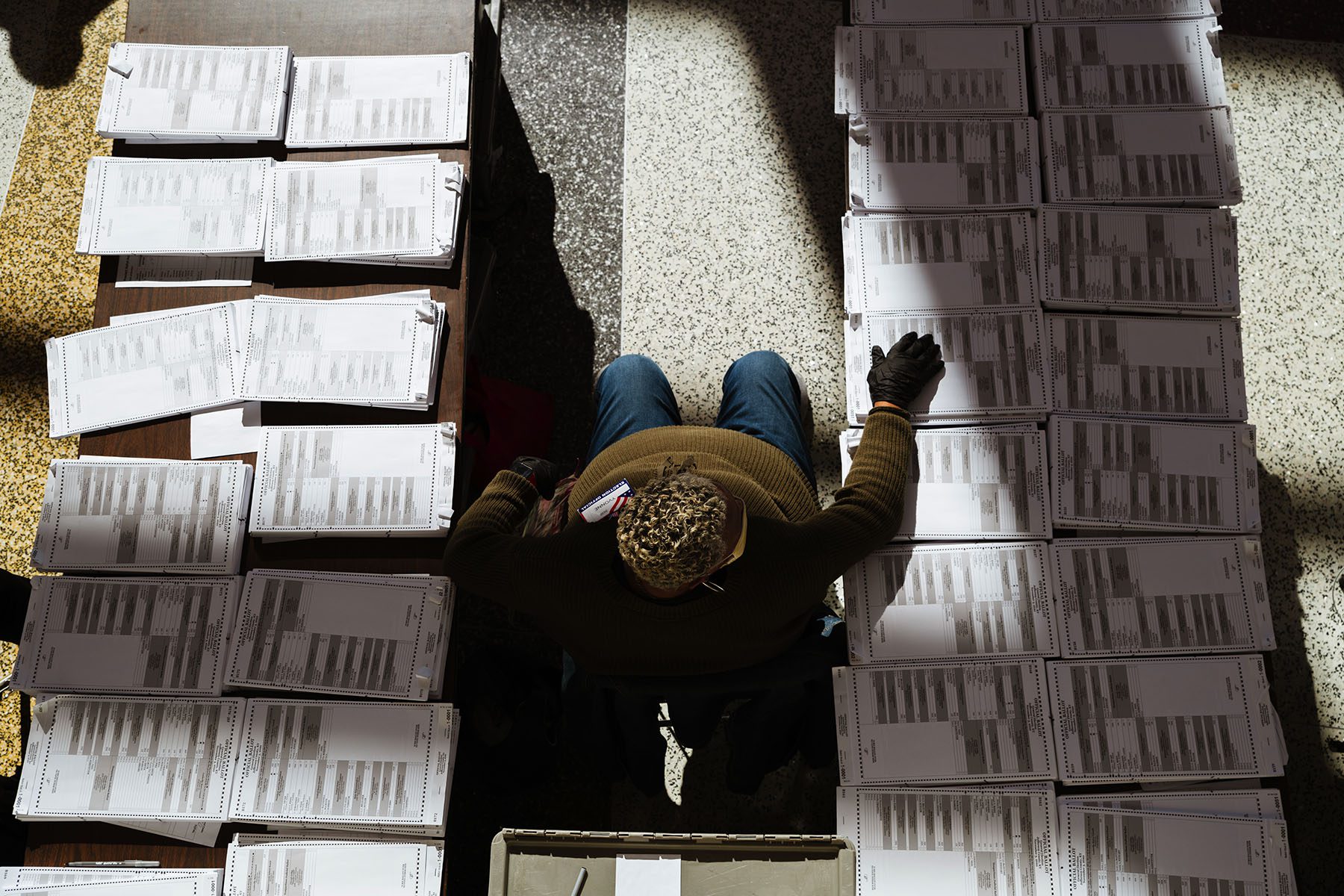 A man sits between two tables on which a laid stacks of blank ballots.