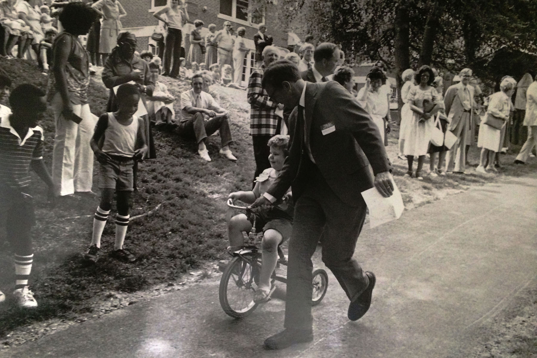 In this black and white image from 1978, a man leads a child on a tricycle down a bike train while people watch, smiling and laughing.