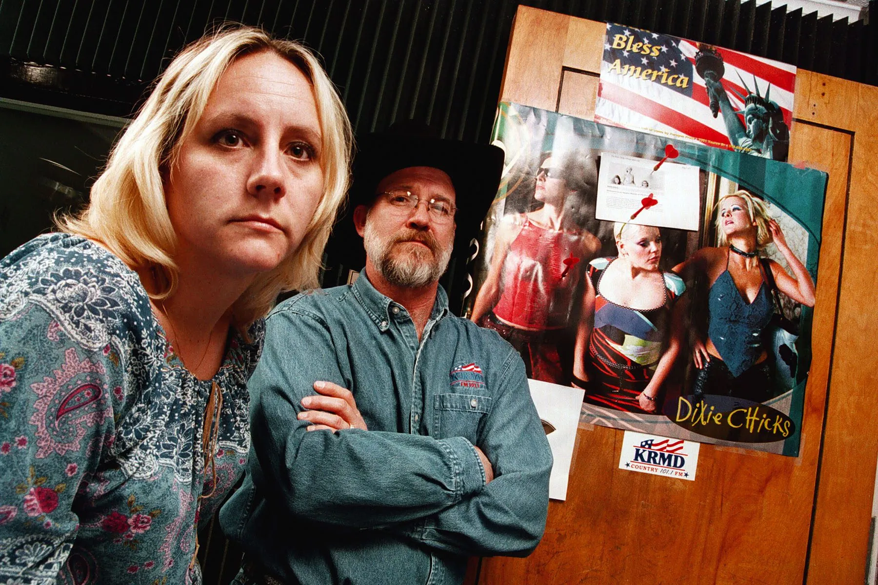 A woman and a man pose for a portrait in a radio studio. Behind them is a poster of The Chicks on which red darts are scattered.