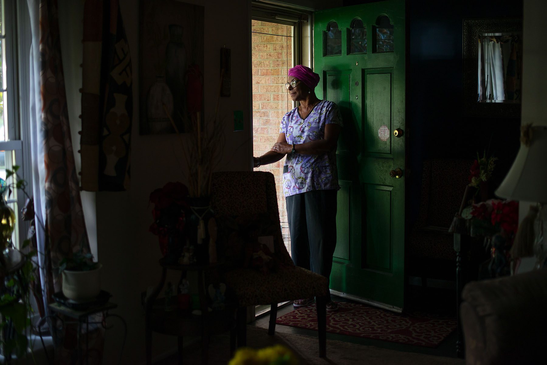 A woman wearing a pink turban and scrubs in seen looking out a screen door in her living room.