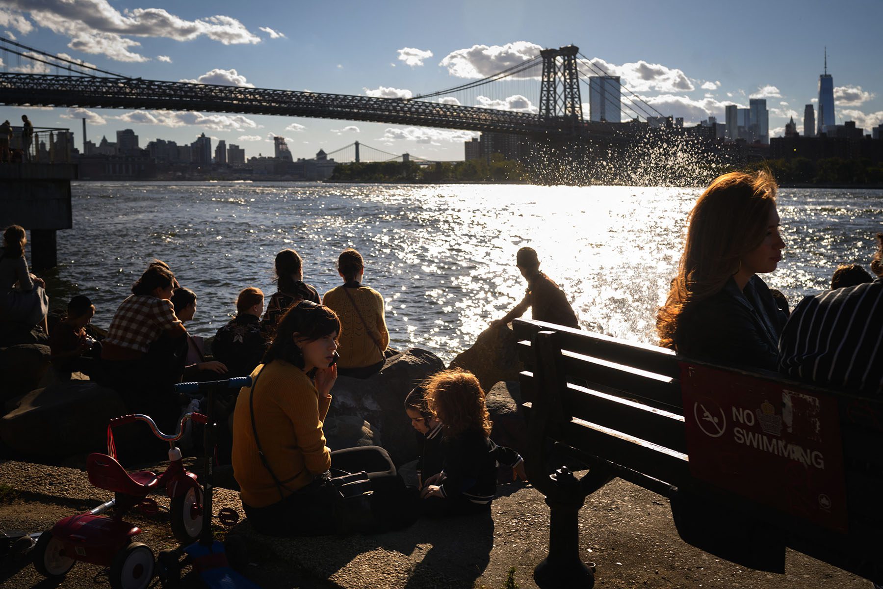 A woman accompanied by two children is on the phone as people sit near the East River. The manhattan skyline is seen in the background.