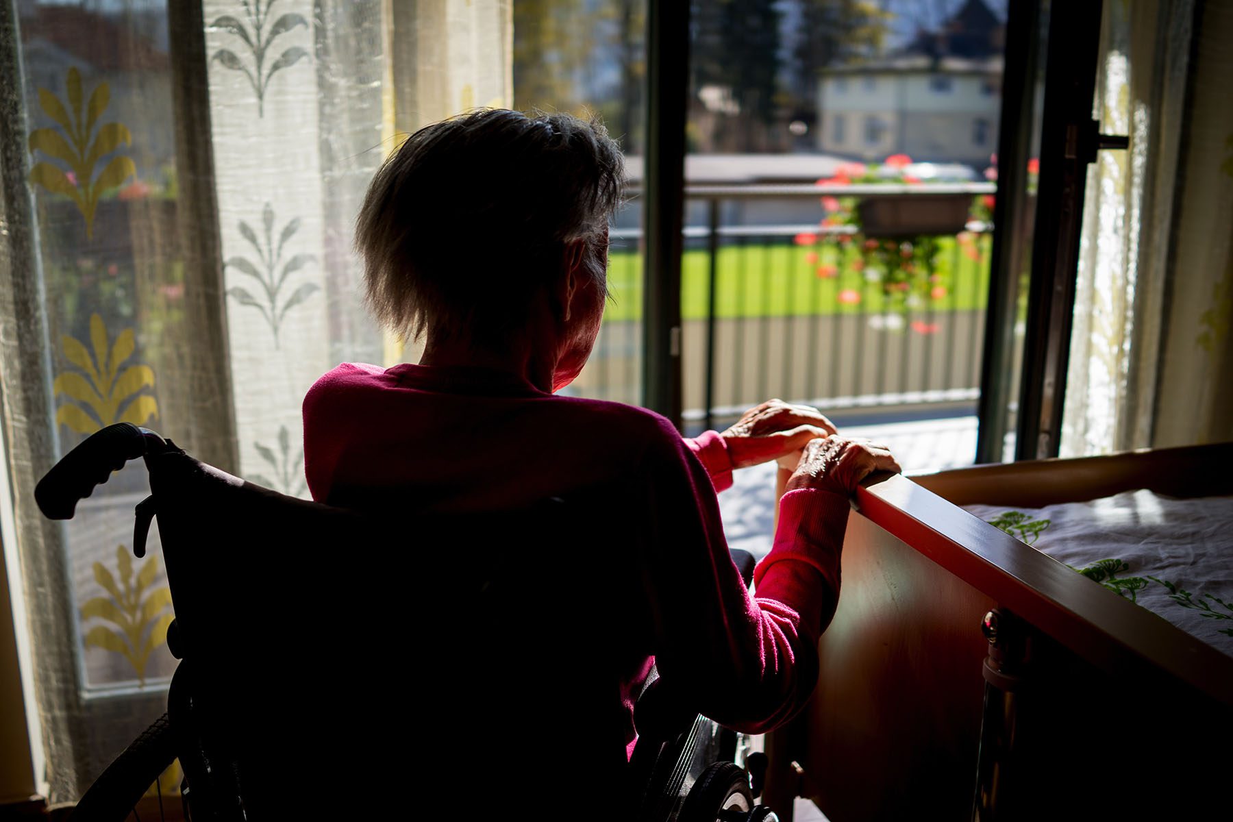 An older person in a wheelchair looks out a glass door. Their face is obscured while the sun shines on their hands.