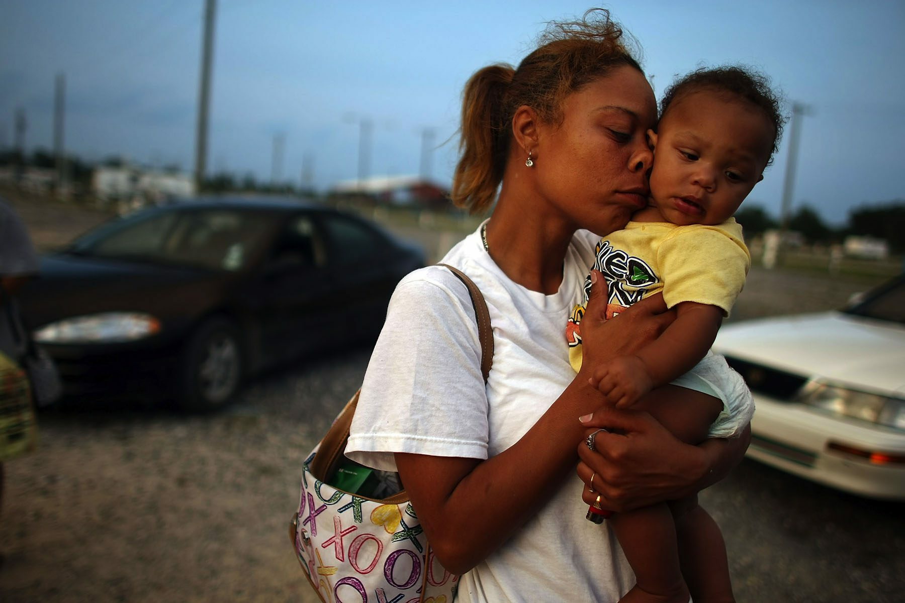 A young woman presses her face against a young child's face while standing on rubble road where cars are parked.