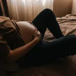A pregnant woman holds her belly while sitting on a bed. Gold light floods the room.