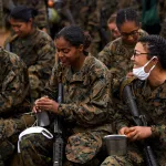Female U.S. Marines eat oranges and speak to each other. They are wearing fatigues and holding rifles.