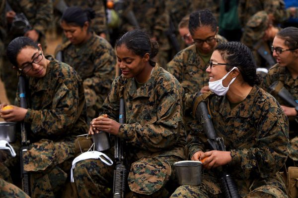 Female U.S. Marines eat oranges and speak to each other. They are wearing fatigues and holding rifles.