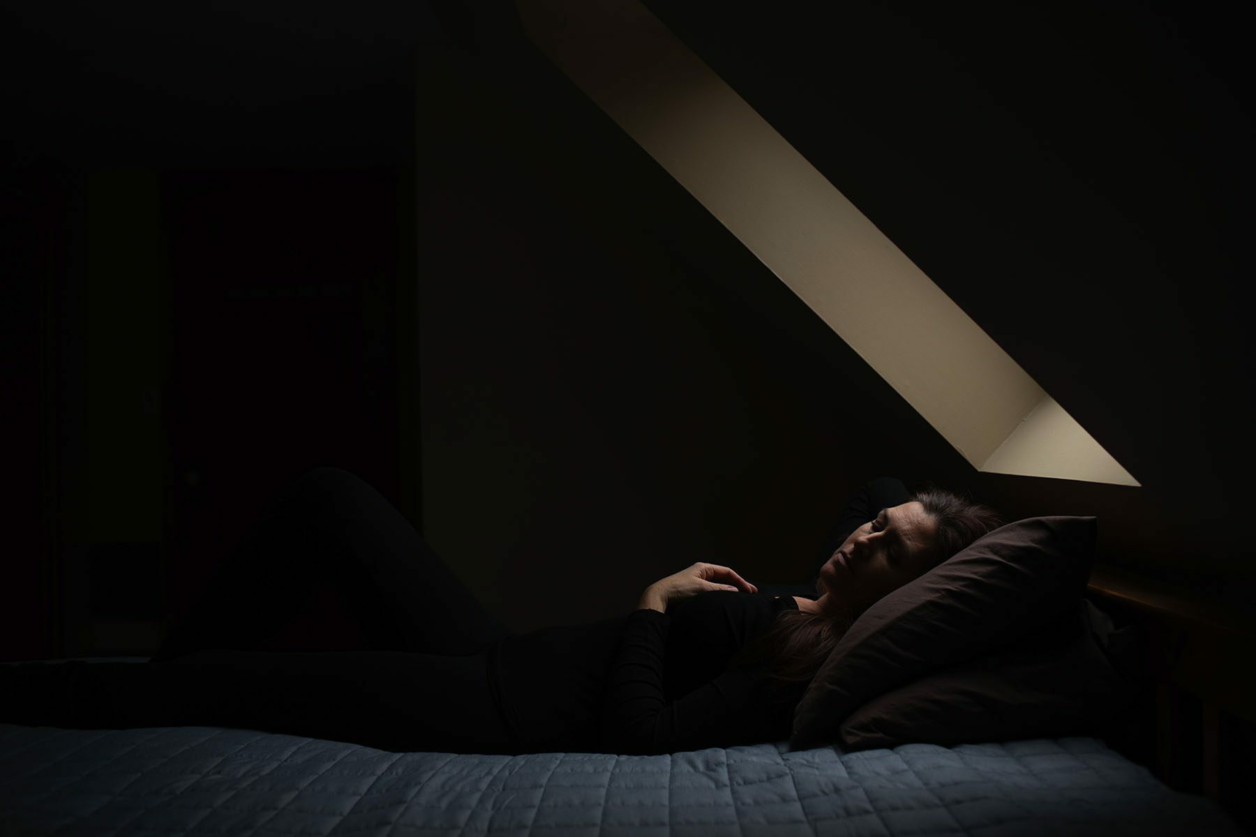 A woman lays on a bed as soft light trickles into the room from a skylight.