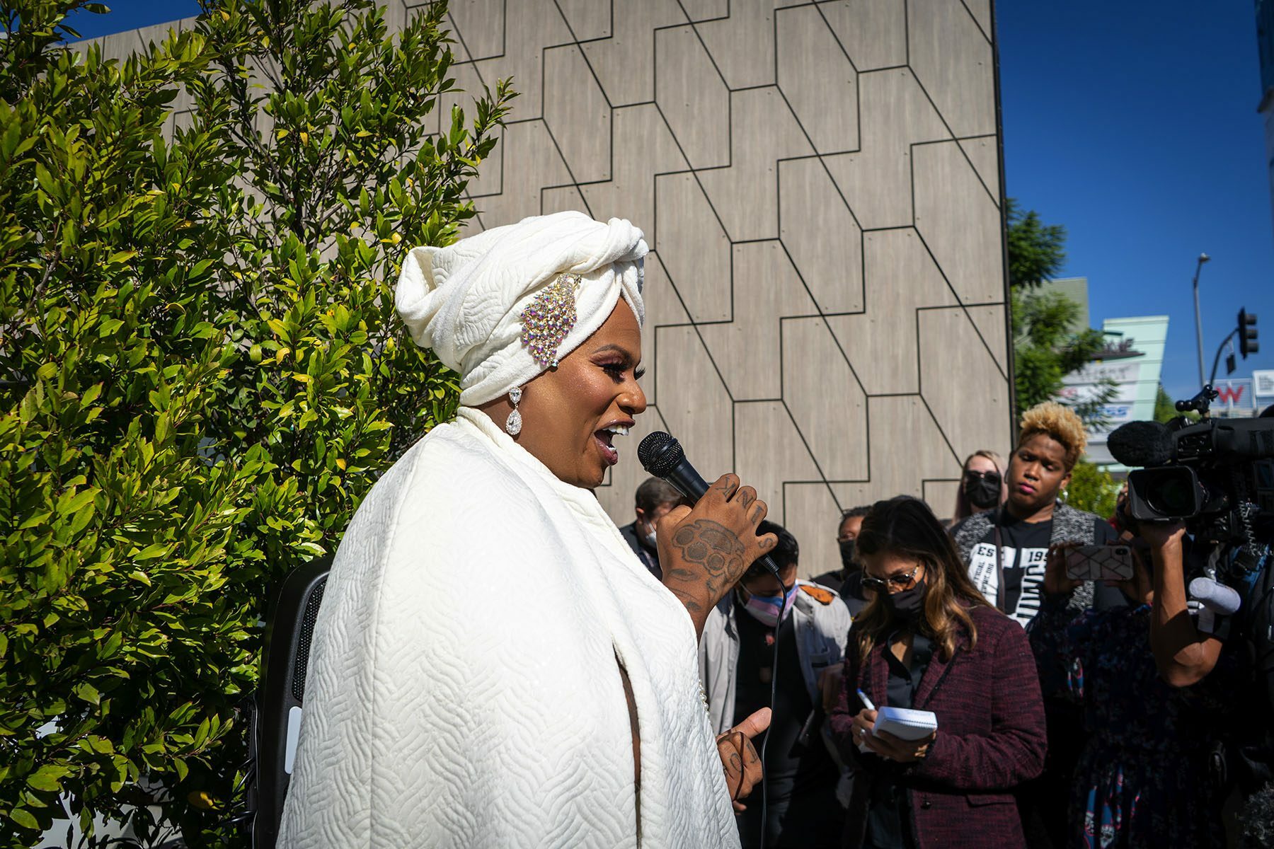 Ashlee Marie Preston dressed in a white cape and a white turban holds a microphone as people and press surround her.