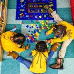 Three little girls seen from above play on a colorful classroom floor.