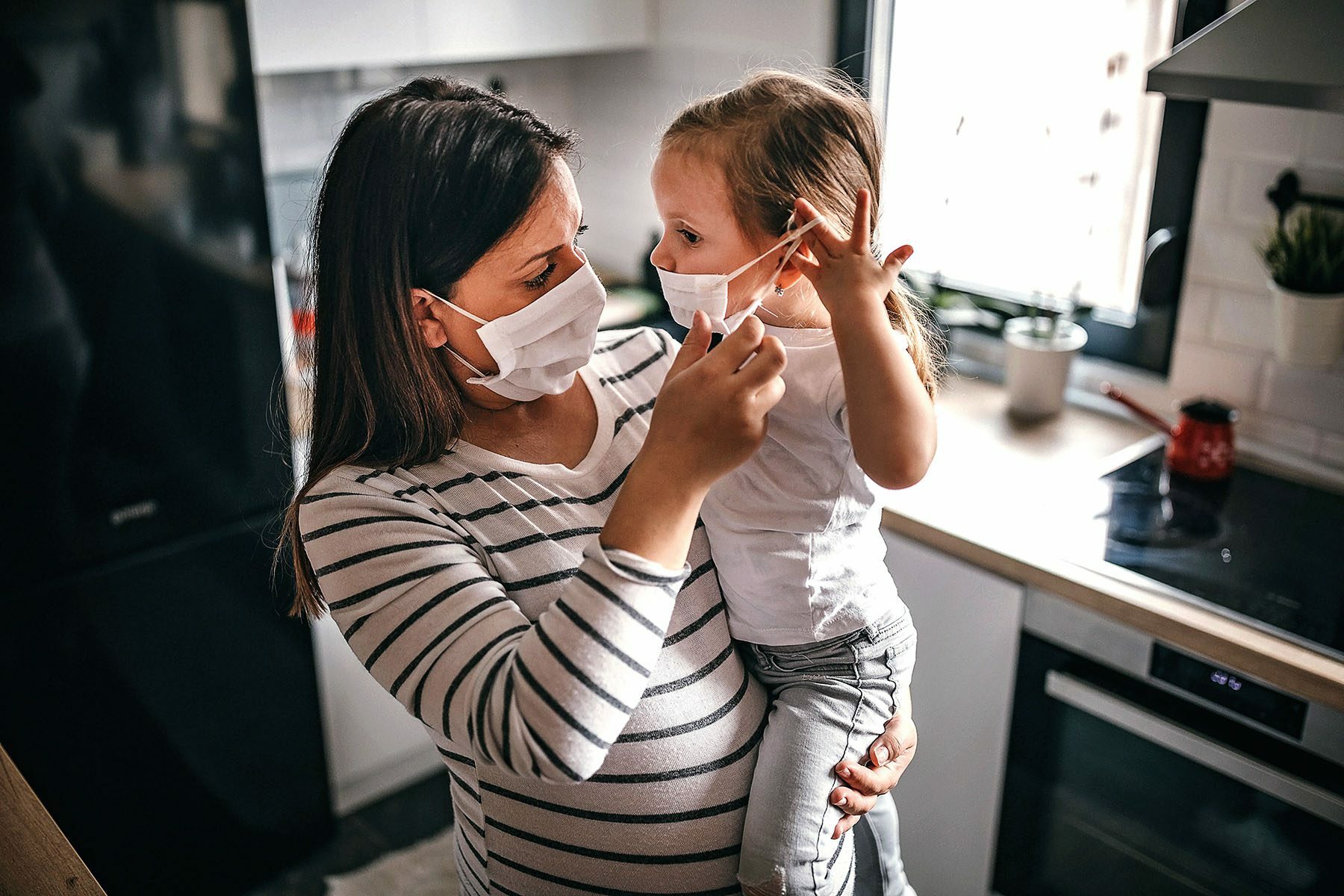 A pregnant woman wearing a face mask helps her young daughter put her face mask on.