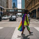A young black child crosses the street wearing a pride flag around their shoulders and a pride bandana around their wrist.