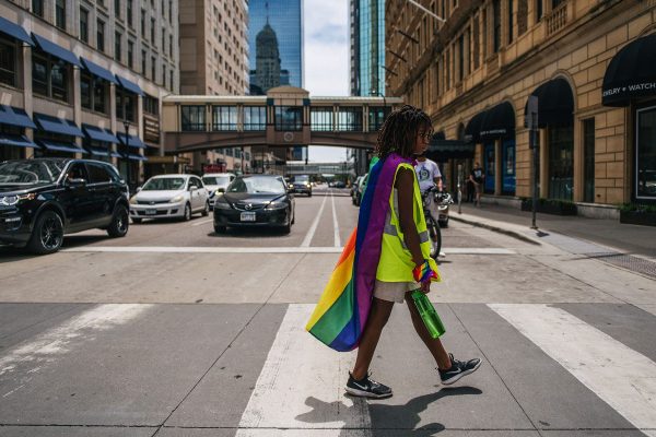 A young black child crosses the street wearing a pride flag around their shoulders and a pride bandana around their wrist.