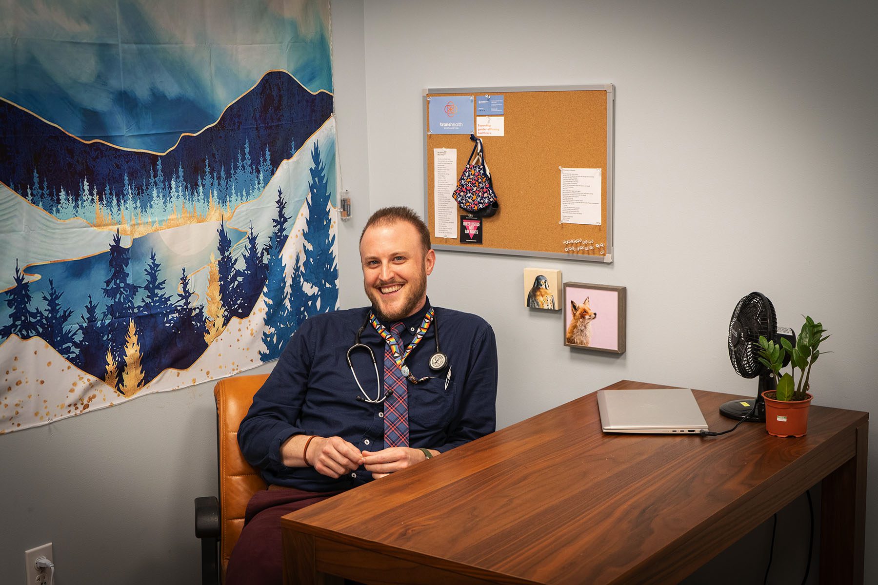 Dr. Sackett-Taylor smiles while sitting at his desk. The room is simply decorated: a corkboard, a plant, a tapestry with blue trees and mountains.