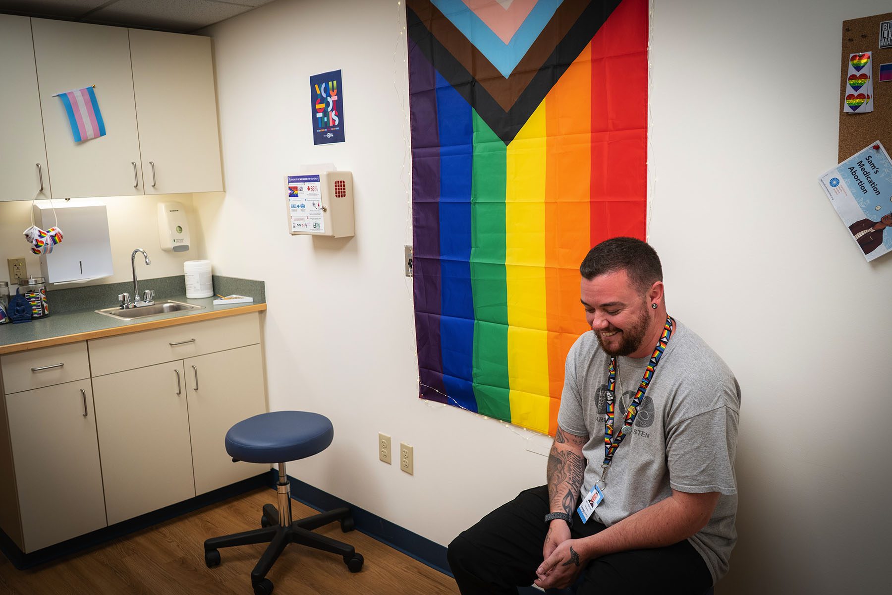 Arin McKona looks down and smiles as he sits on a stool in an examination room draped with a progressive pride flag.
