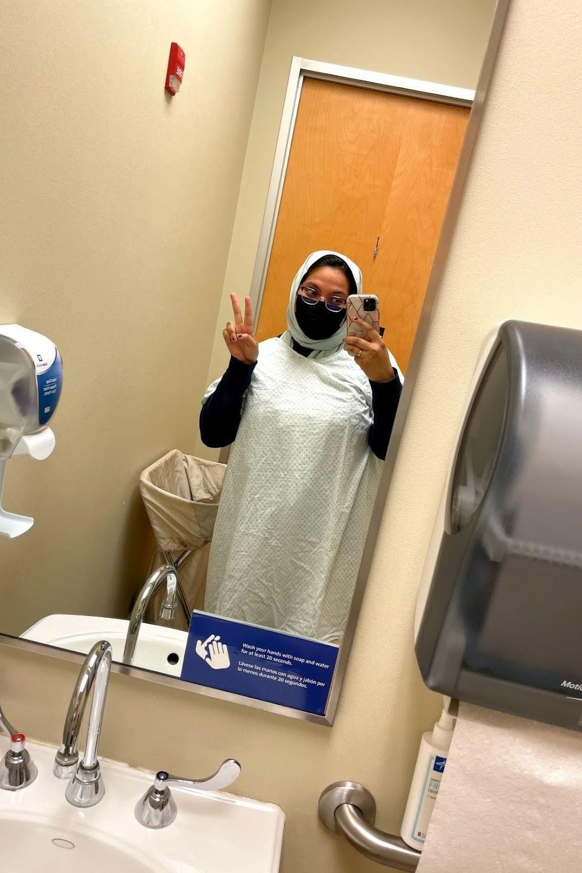 a mirror selfie of Nira Allam flashing a peace sign and wearing a patient gown.