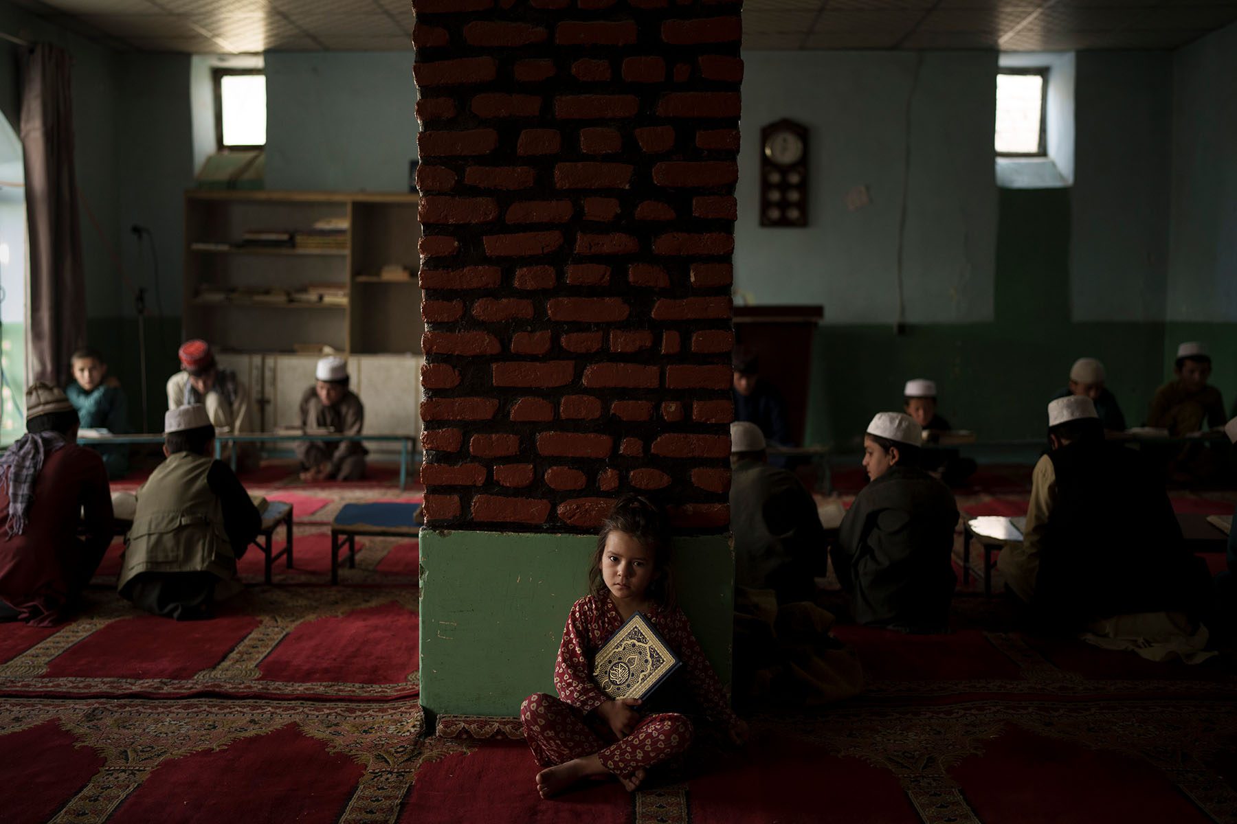 A little girl sits alone and holds a Quran in a classroom.